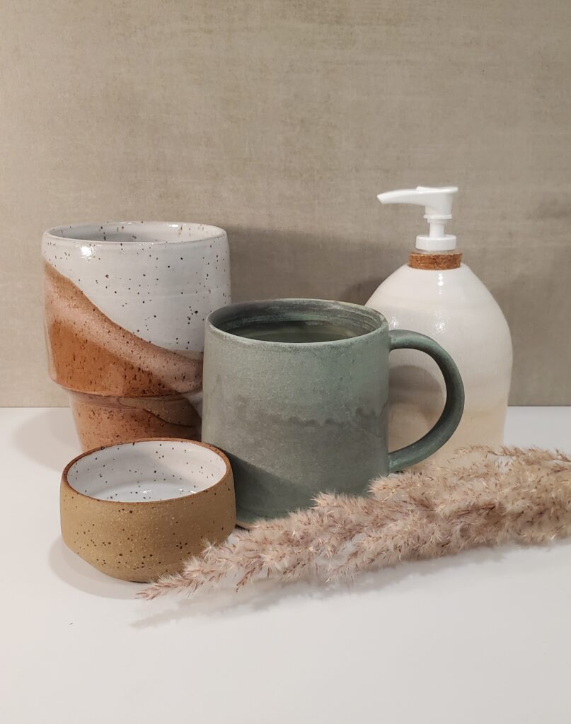 collection of functional pottery pieces in muted glaze colors including a large tumbler, a coffee mug, a small bowl and a ceramic soap dispenser